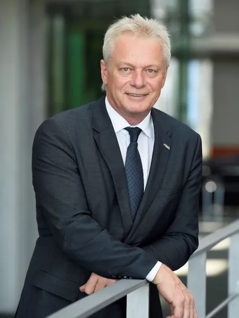 Portrait of Alois Rainer in a dark suit, smiling and leaning on a metal railing in a modern building