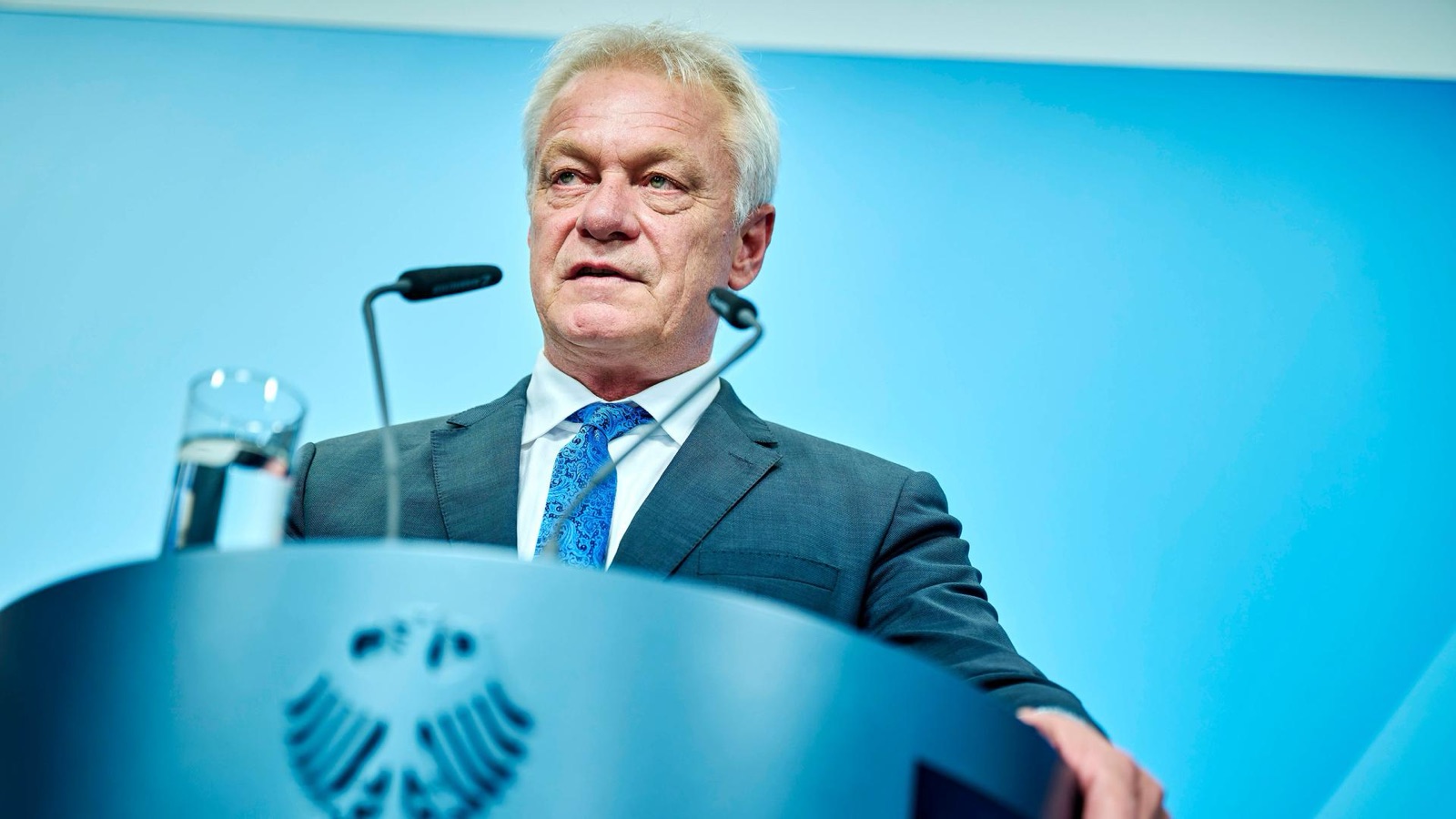 Alois Rainer speaking at a lectern bearing the federal eagle, against a pale blue background